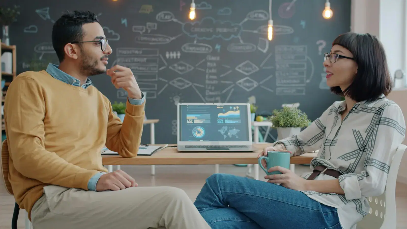 Two professionals having a discussion in a modern office with data charts on a screen.